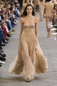 Runway model in a cream, tiered, sheer gown with a jeweled bodice walking the catwalk as seated audience watch.