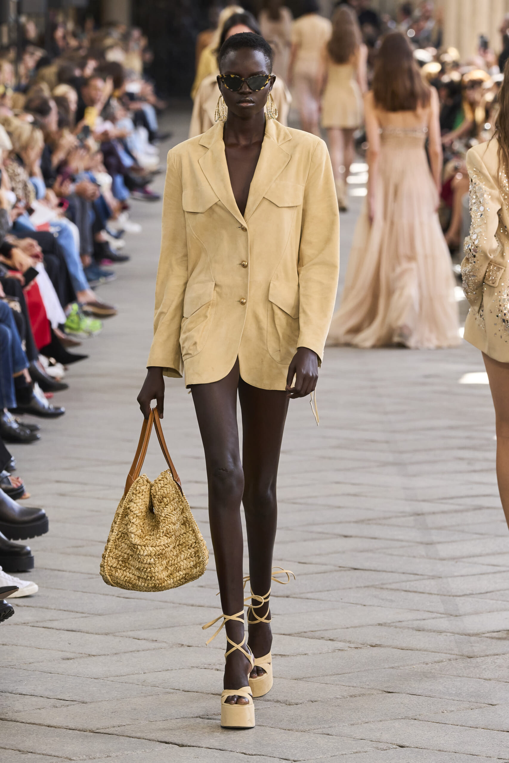 Model walks a fashion show runway wearing a tan blazer, oversized sunglasses, and holding a woven straw bag with platform sandals.