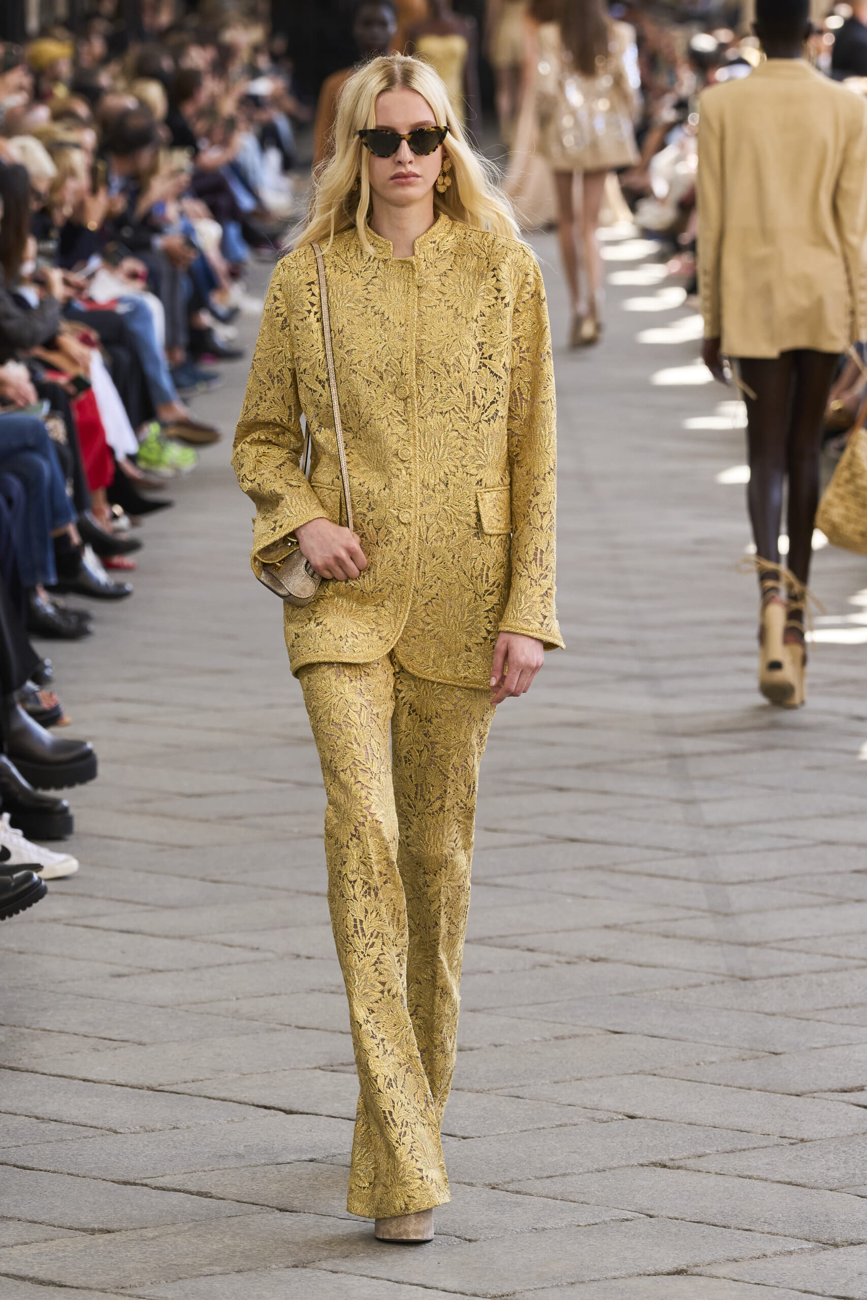 Model walking the runway in a gold lace suit with sunglasses, audience watching the show along the sides.