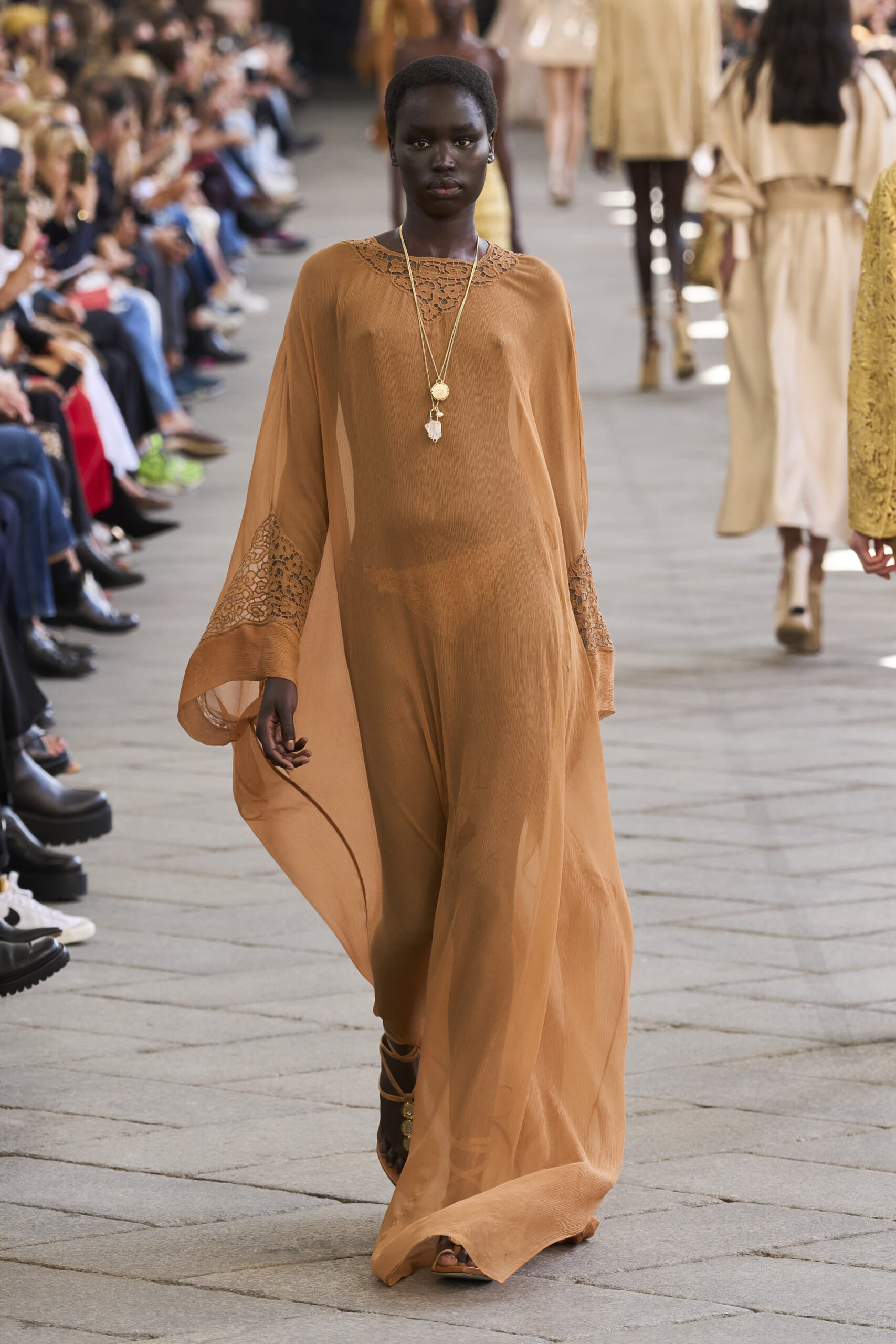 Model walking a fashion show runway in a long peach dress with lace details and layered necklaces, audience visible on both sides.