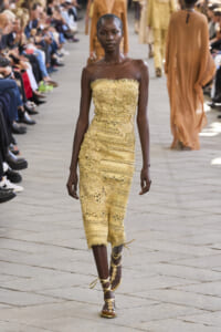 Model walks runway in a gold strapless lace dress with gold gladiator sandals; seated audience along the sides.