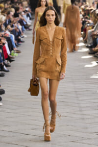 Model walks the runway in a tan suede lace-up mini dress, carrying a brown handbag and wearing matching platform sandals.