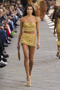 Model walks a gold crochet strapless two-piece outfit on an outdoor fashion show runway with seated spectators along the sides.