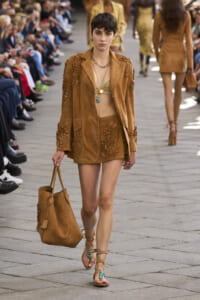 Model walks a runway in a tan suede suit with floral appliqués, turquoise necklace, and matching tan bag.