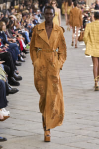 Model on a runway wearing a tan suede coat with floral appliqué and a matching belt, audience on both sides watching