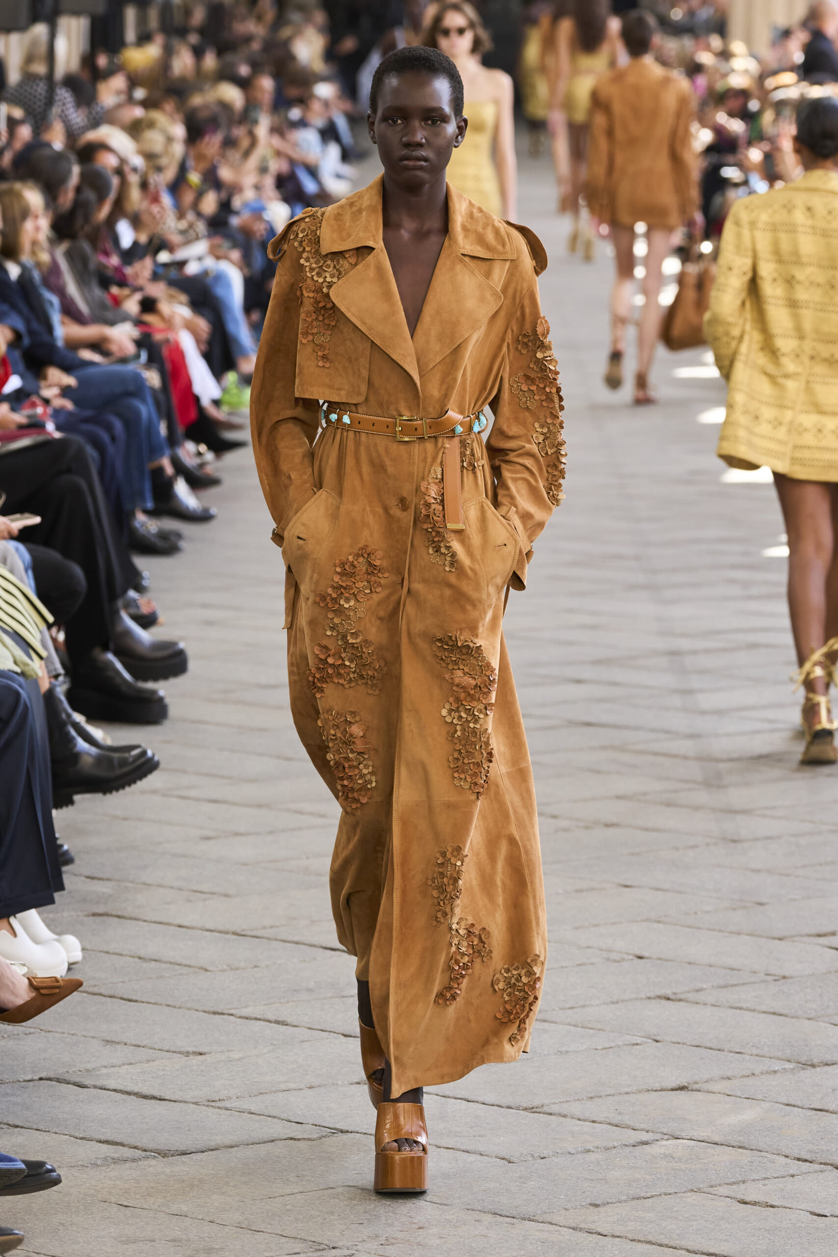 Model on a runway wearing a tan suede coat with floral appliqué and a matching belt, audience on both sides watching