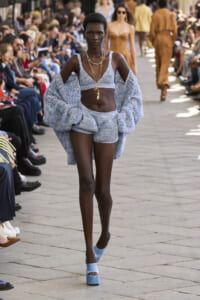 Runway model in a light blue crochet two-piece with cape, walking the catwalk as spectators watch.