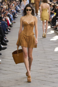 Model walking an outdoor fashion show in a strapless caramel dress with a large brown tote and platform sandals, wearing chunky sunglasses and gold earrings.
