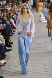 Model walks the fashion runway in a sheer light-blue blouse with blue embroidery and light-wash denim jeans, audience along the catwalk.