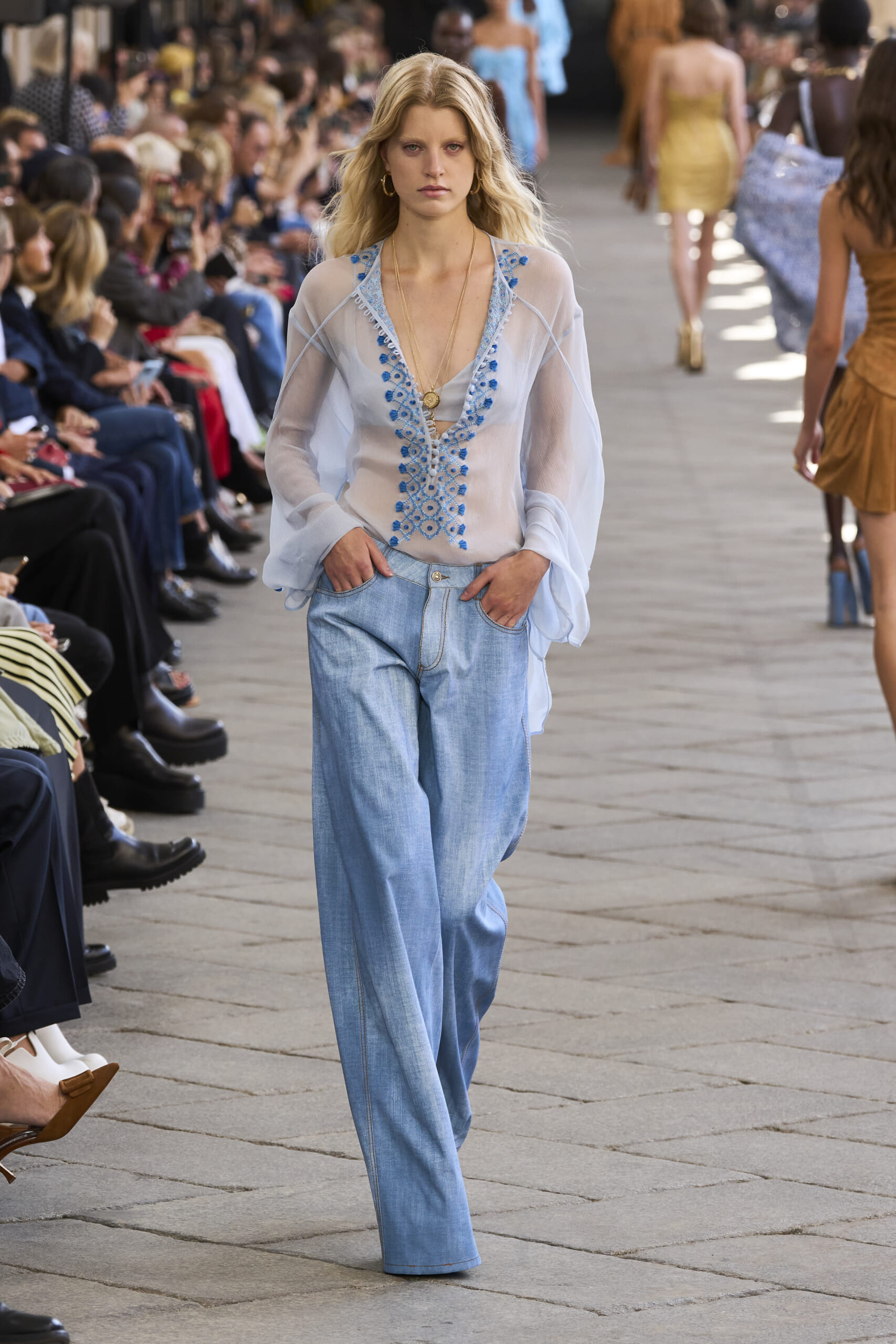 Model walks the fashion runway in a sheer light-blue blouse with blue embroidery and light-wash denim jeans, audience along the catwalk.