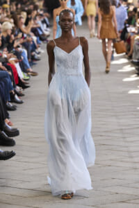 Model walking a runway in a pale blue, pleated gown as spectators watch the show on both sides of the catwalk.
