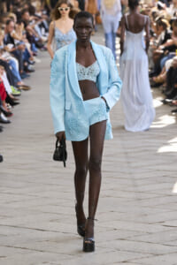 Model walks the runway in a light blue suit with lace top, surrounded by an audience at a fashion show.