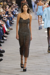 Model walks down a runway in a glittery sheer black dress with lace bodice, audience on both sides.