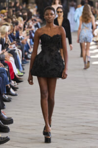 Model walking a black floral-embellished strapless dress on a runway with seated audience on both sides, foreground focus on the dress.
