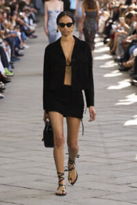 Model walks a fashion runway in a black jacket and mini skirt, wearing sunglasses and gold jewelry, with spectators on both sides.