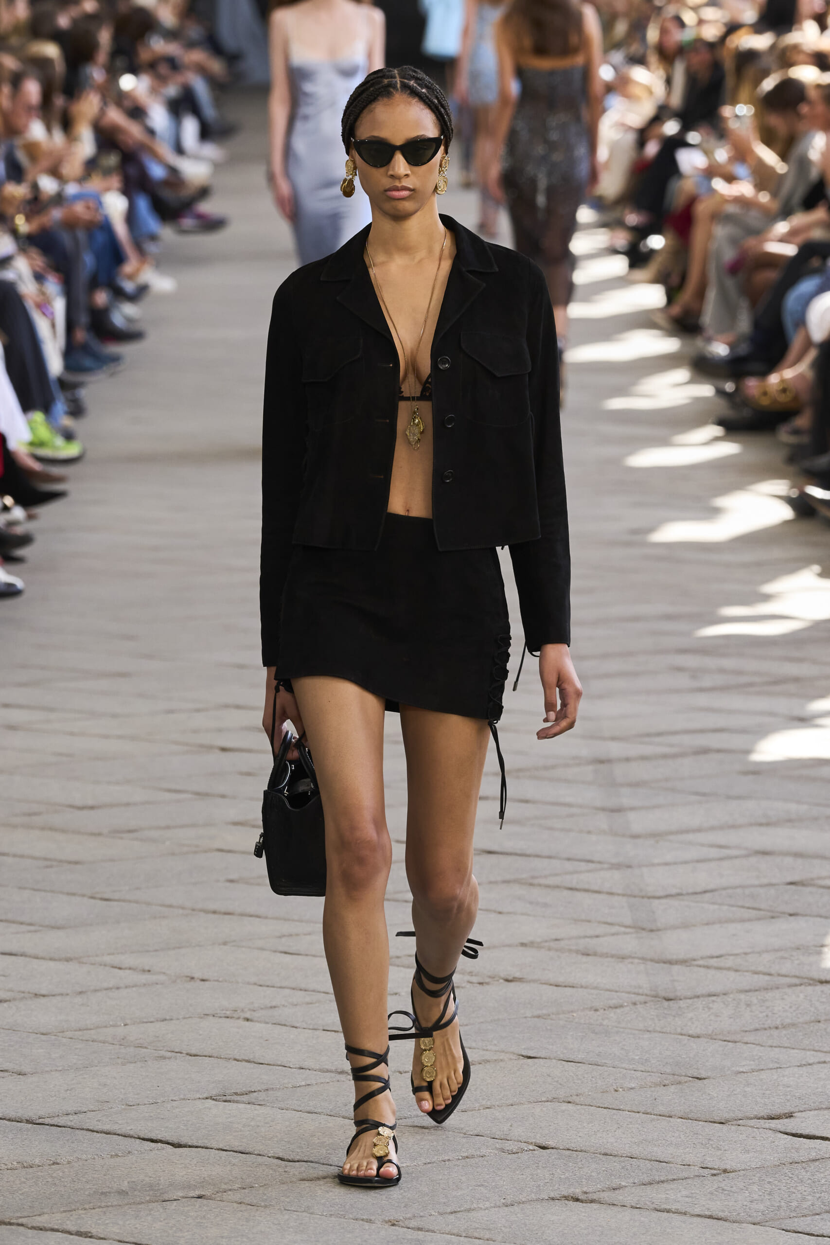 Model walks a fashion runway in a black jacket and mini skirt, wearing sunglasses and gold jewelry, with spectators on both sides.