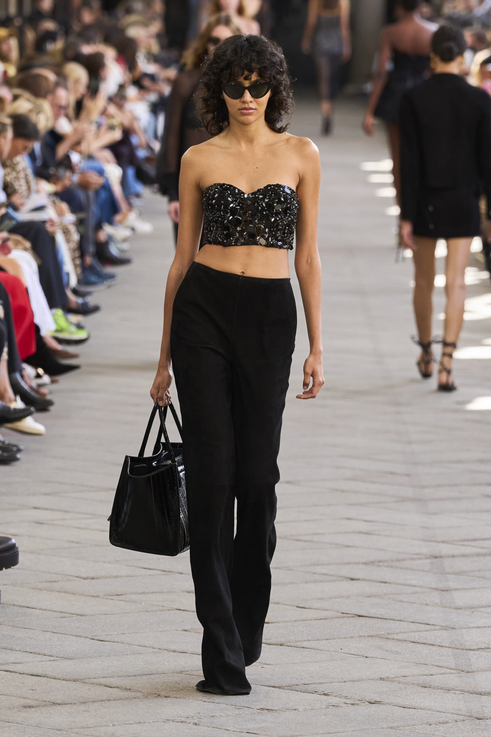 Model walking a fashion runway in a black sequined strapless top and black pants, holding a black handbag with sunglasses on.