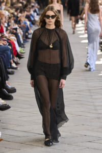 Model walks a runway in a sheer black outfit with cape, wearing sunglasses and gold pendant necklaces, flanked by seated audience members.