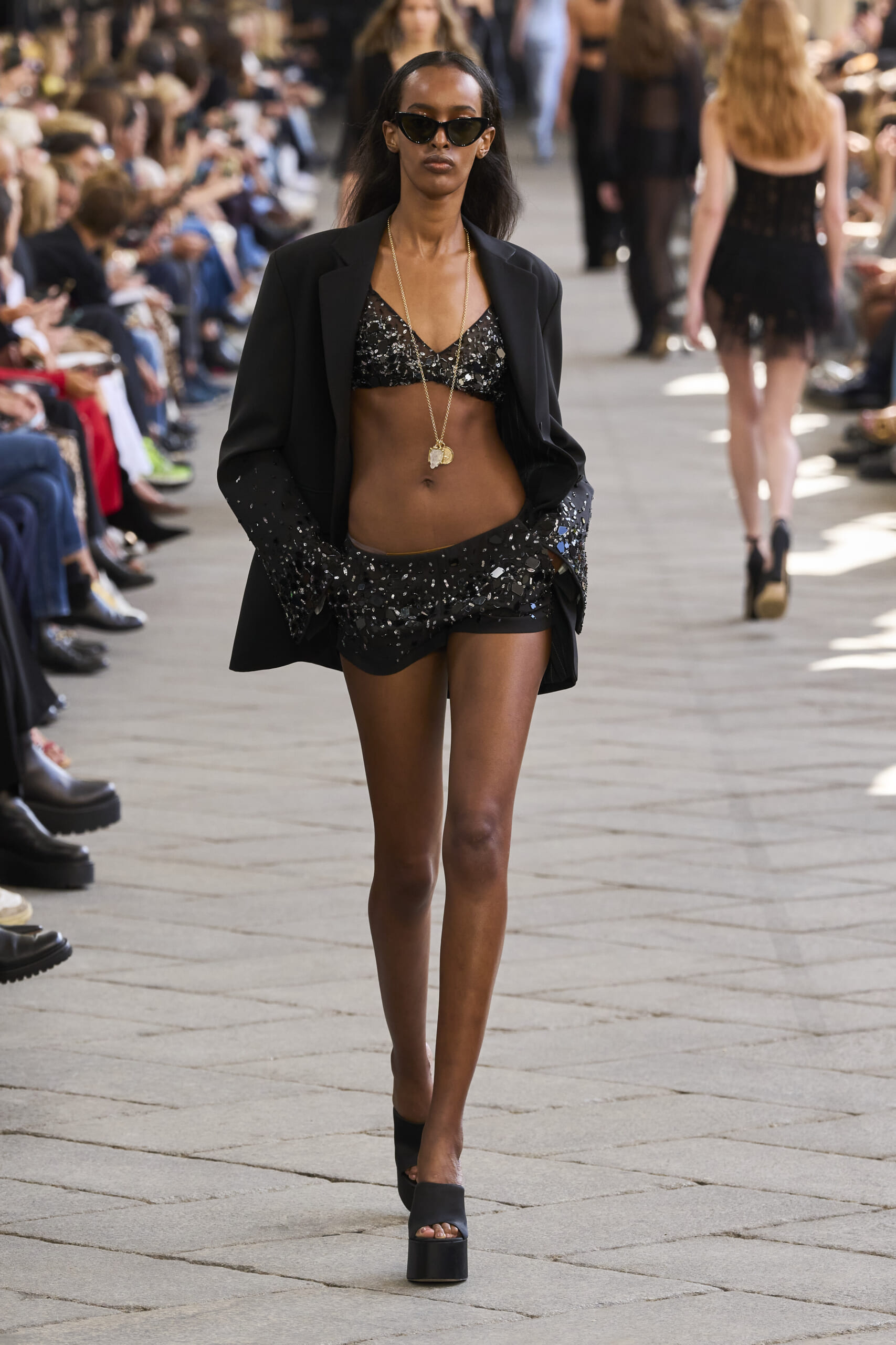Model walks runway in a black sequin crop top, high-waisted shorts, and blazer with sunglasses and a long gold necklace.
