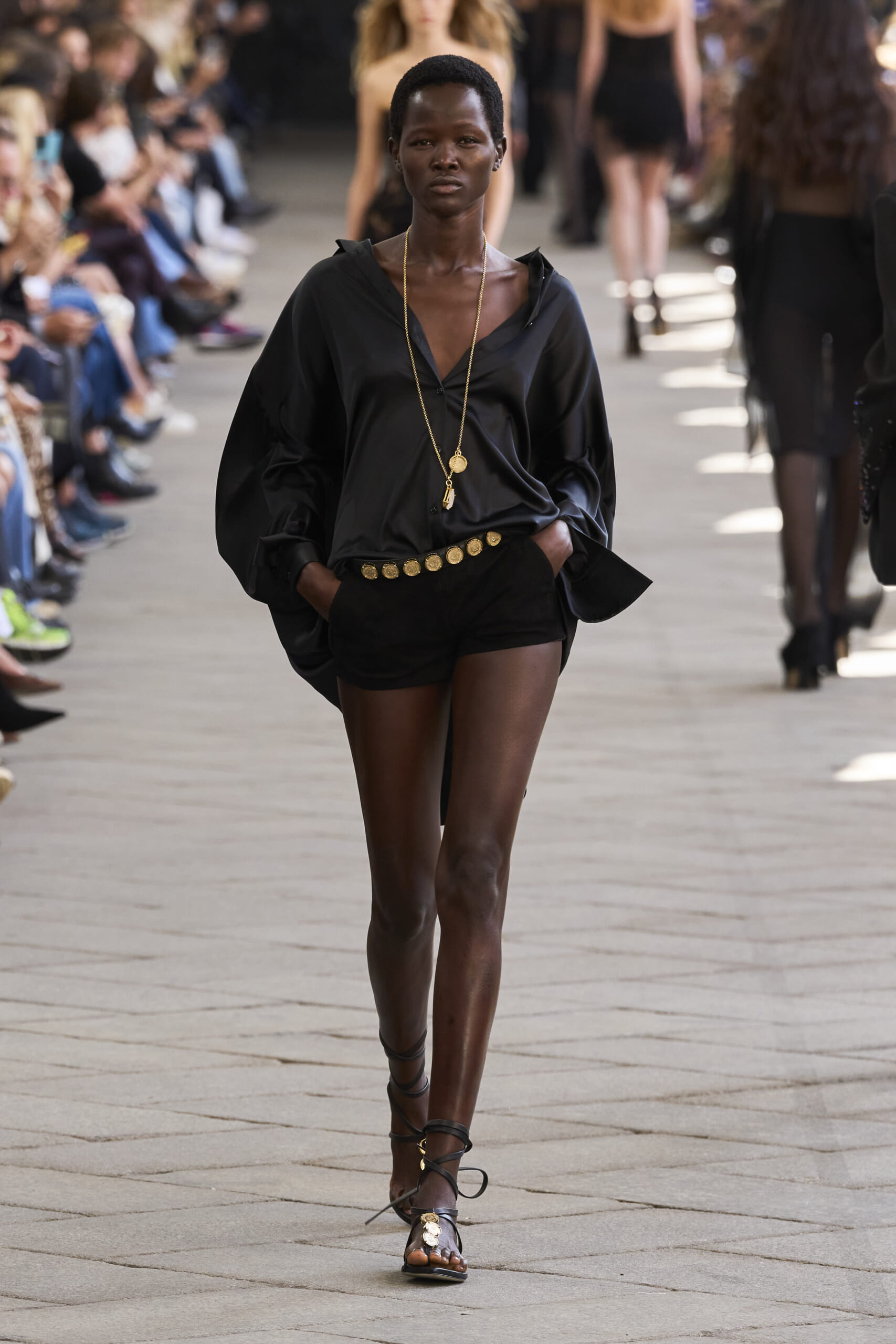 Model walking a fashion runway in a black blouse with a gold belt and long gold necklace; spectators blur in the background