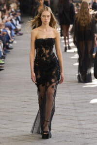 Model walks a runway in a black lace strapless gown with floral appliqués, audience blurred in the background.