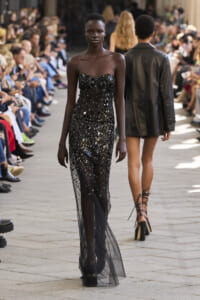 Model walking a fashion show runway in a strapless black sequin gown, audience seated along the sides.
