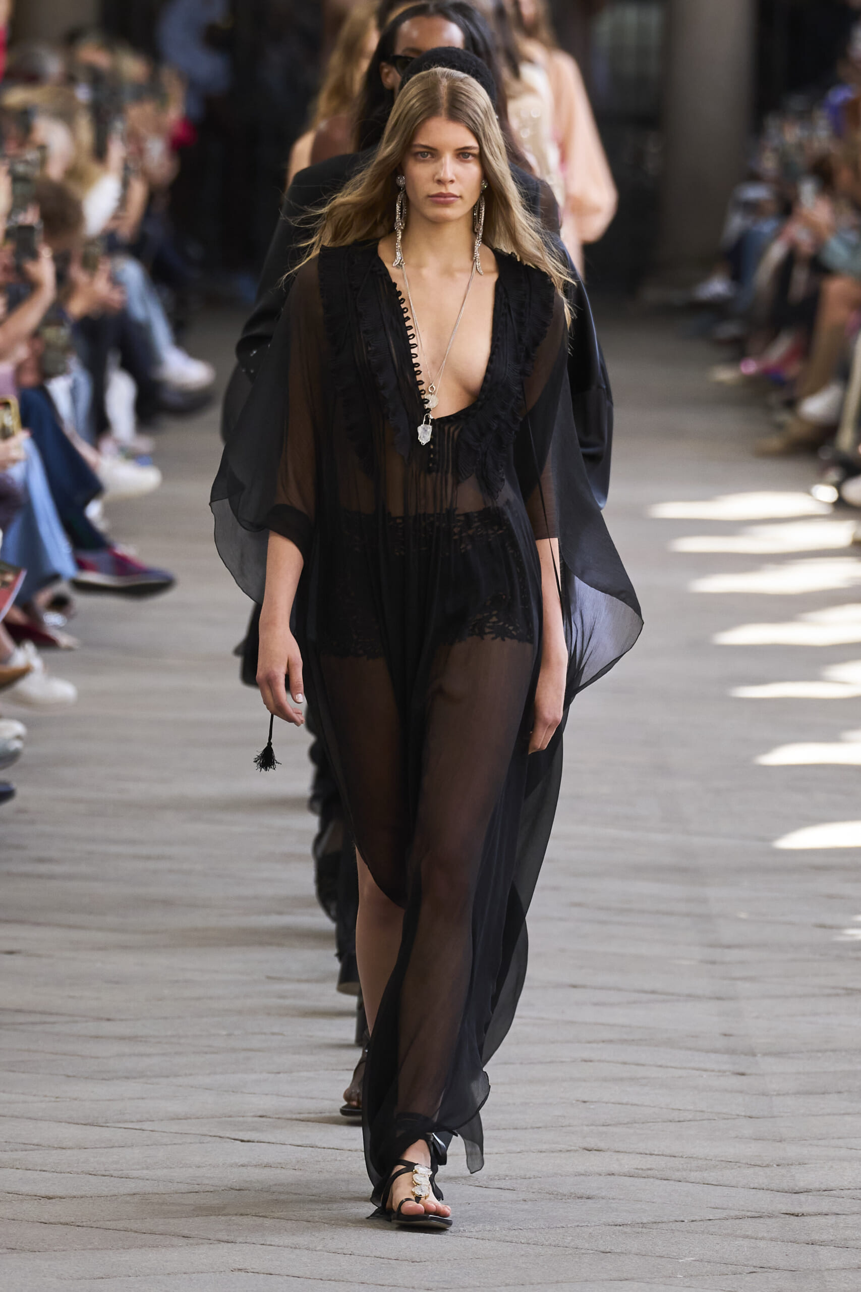 Model walks a fashion show in a sheer black dress with a deep neckline and long layered jewelry, audience on both sides Shaw