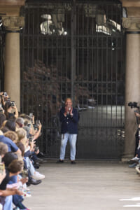 Man in a navy blazer stands in front of ornate gates, clapping to a cheering audience taking photos.