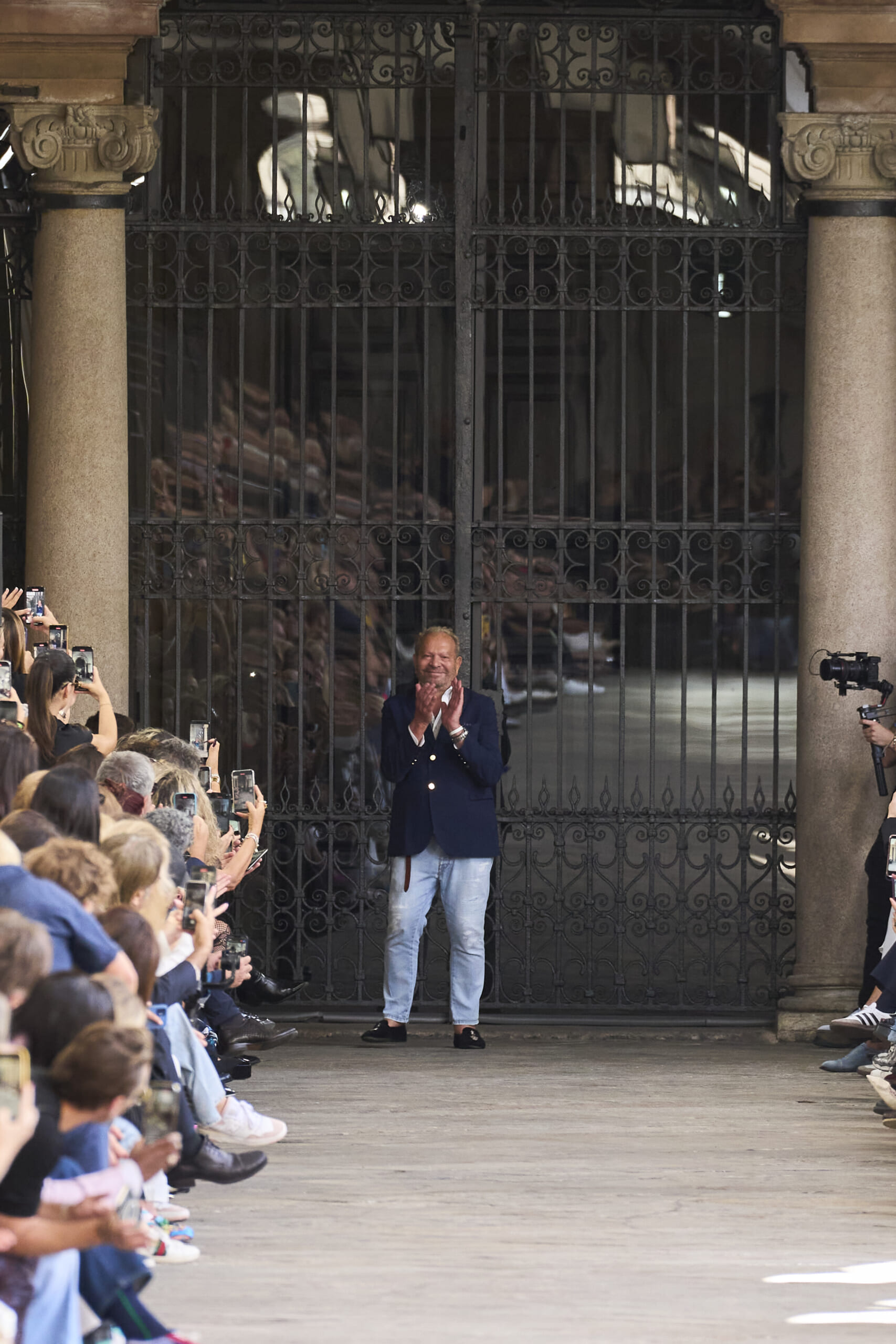 Man in a navy blazer stands in front of ornate gates, clapping to a cheering audience taking photos.