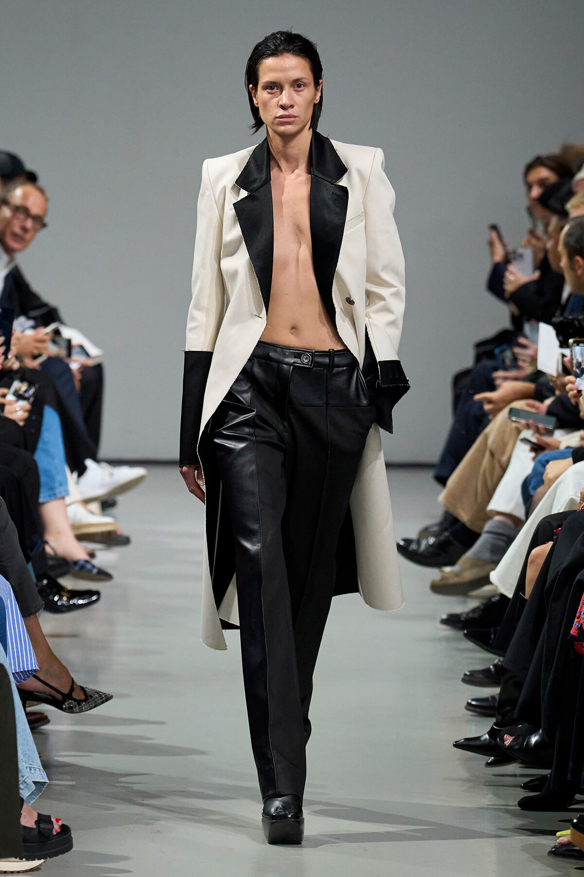 Runway model in a white blazer with black lapels, black leather pants, and visible midriff walking a fashion show; audience on both sides.
