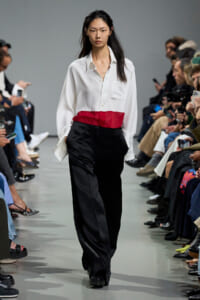 Model on a runway wearing a white button-down shirt with a red waist band and black trousers, audience on both sides watching.