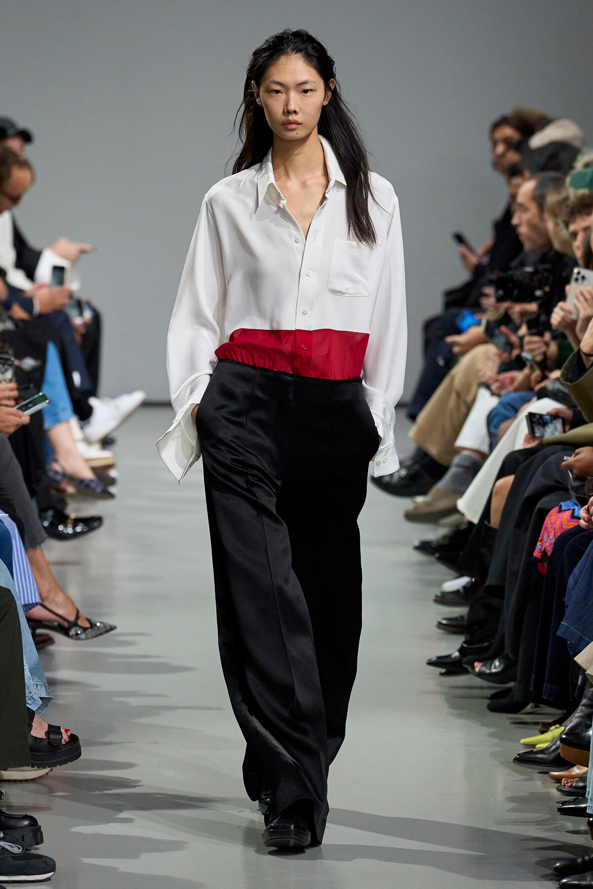 Model on a runway wearing a white button-down shirt with a red waist band and black trousers, audience on both sides watching.
