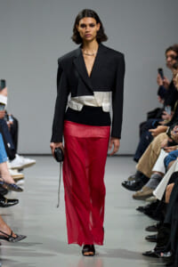 Model on a runway wearing a black blazer with a silver waist belt and sheer red pants, holding a small black clutch while audience watches.