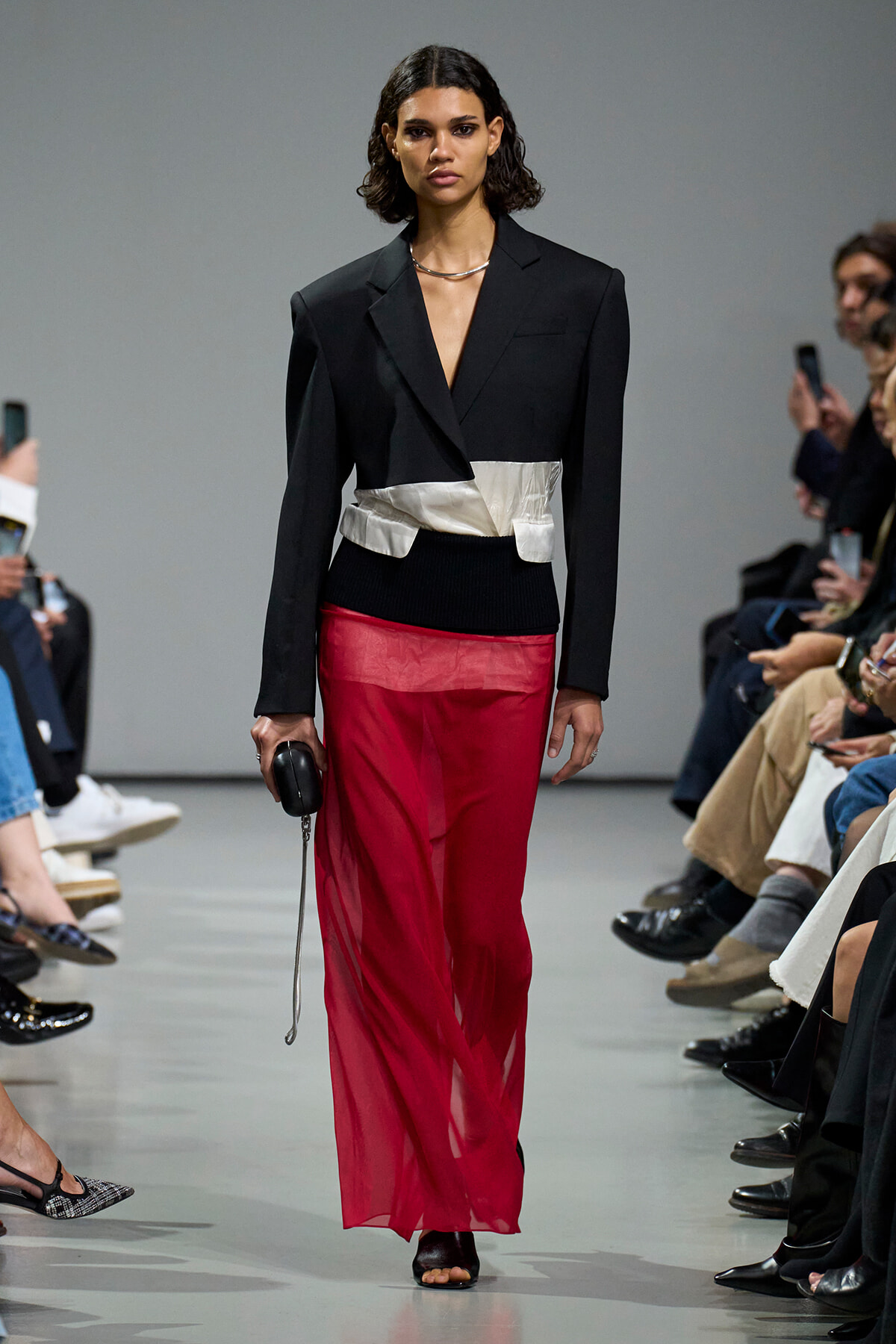 Model on a runway wearing a black blazer with a silver waist belt and sheer red pants, holding a small black clutch while audience watches.