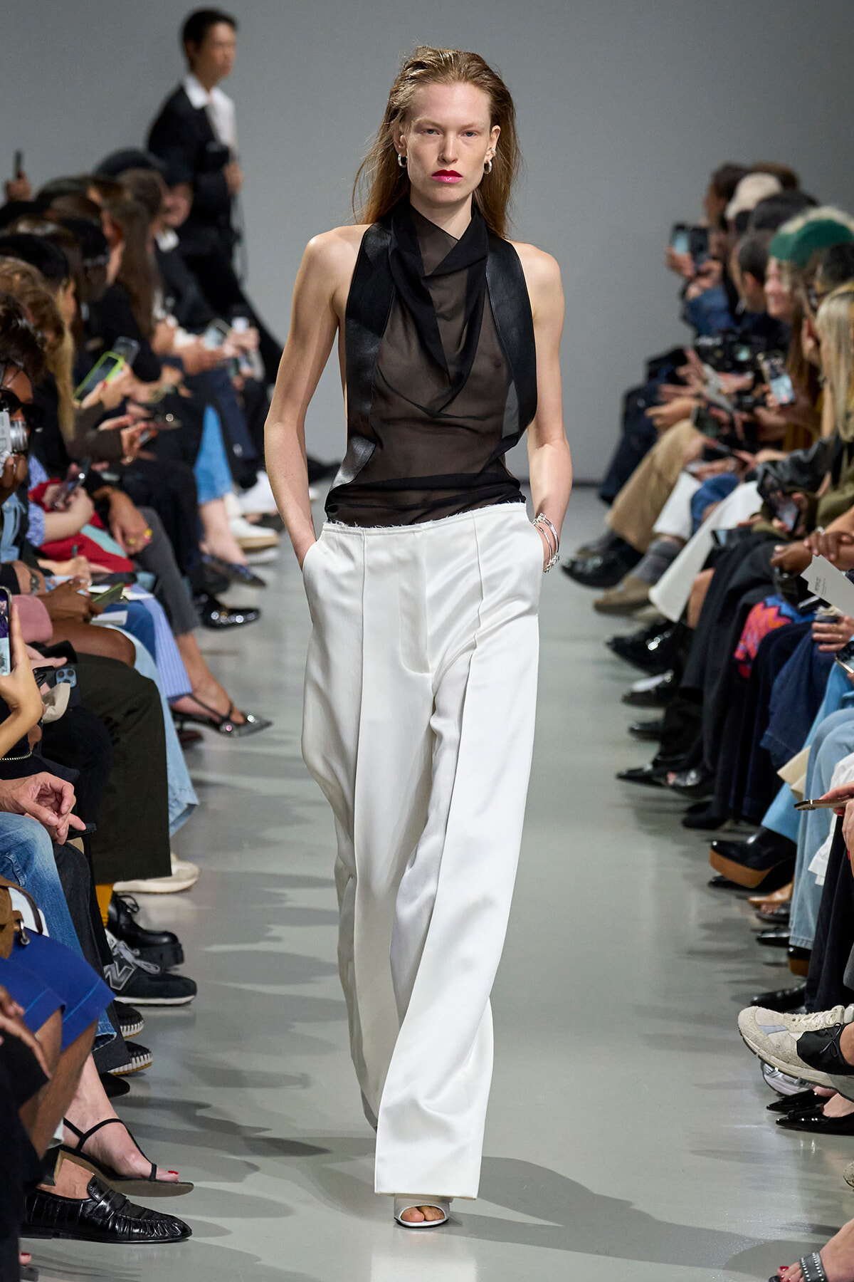 Model walks runway in a black sheer sleeveless top with draped scarf over white wide-leg pants, audience on both sides watching closely.