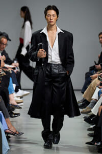 Model walking a fashion show in a black suit with a white shirt, carrying a black clutch bag.