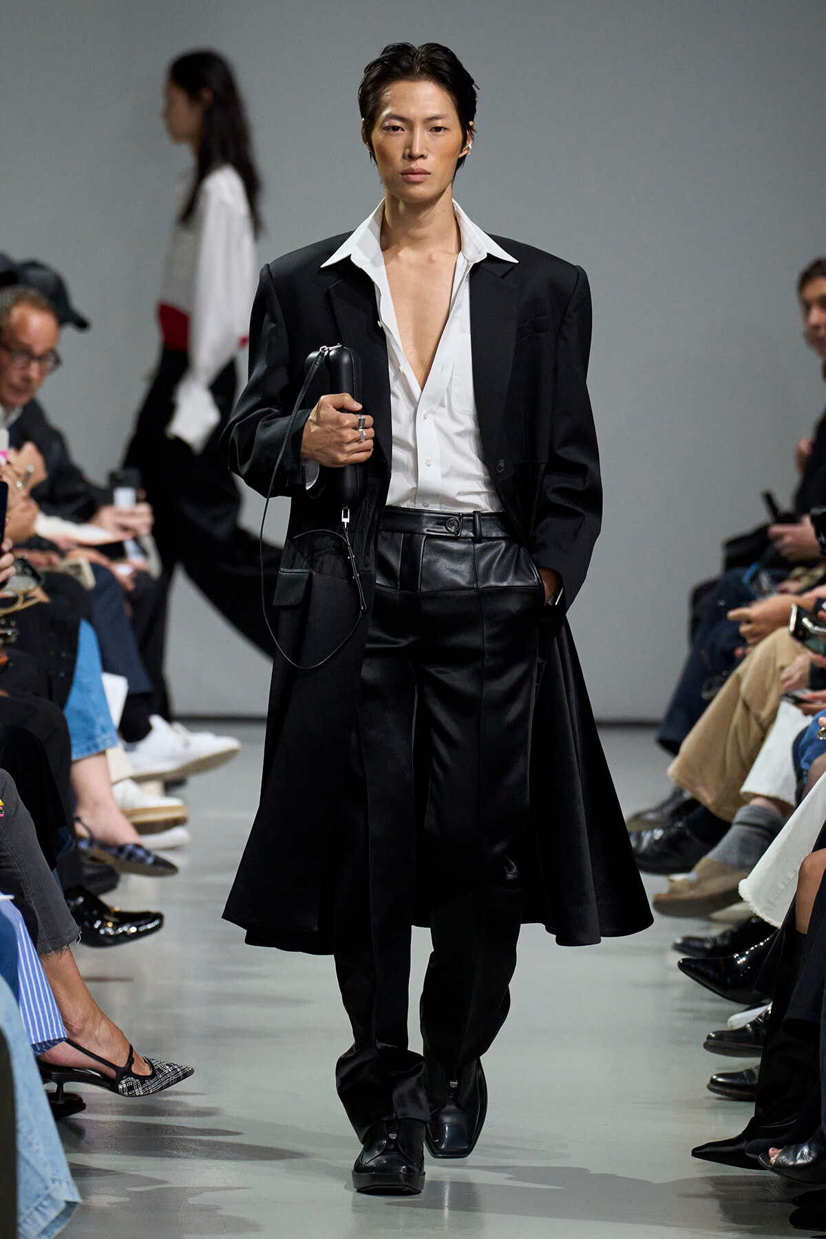 Model walking a fashion show in a black suit with a white shirt, carrying a black clutch bag.
