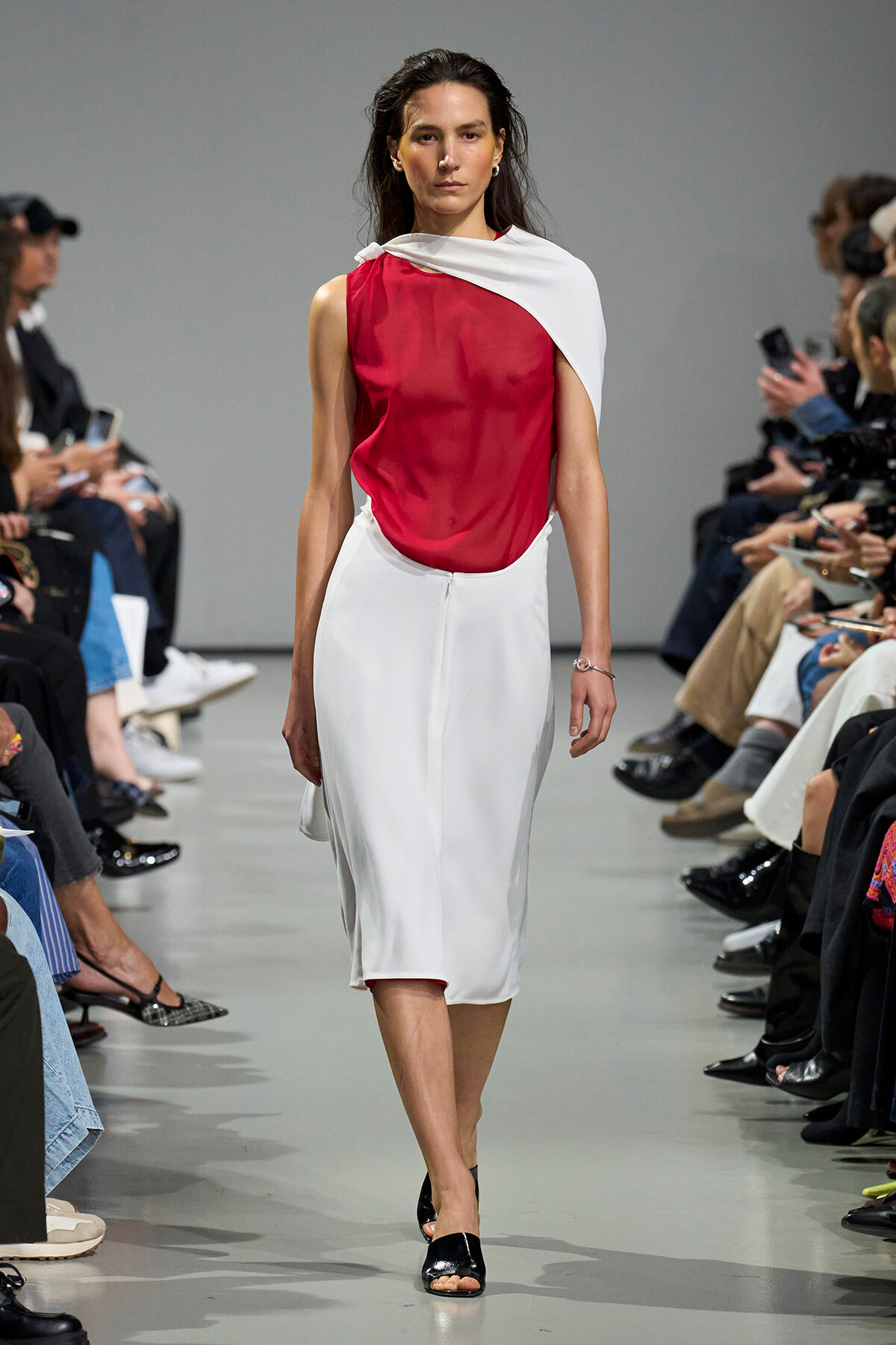 Model walks the runway in a red sleeveless top with a white draped scarf and a white midi skirt, black sandals visible at feet.