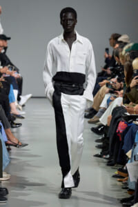 Model walks the runway in a white shirt with a black waist panel and two-tone white/black pants at a fashion show, audience on both sides.