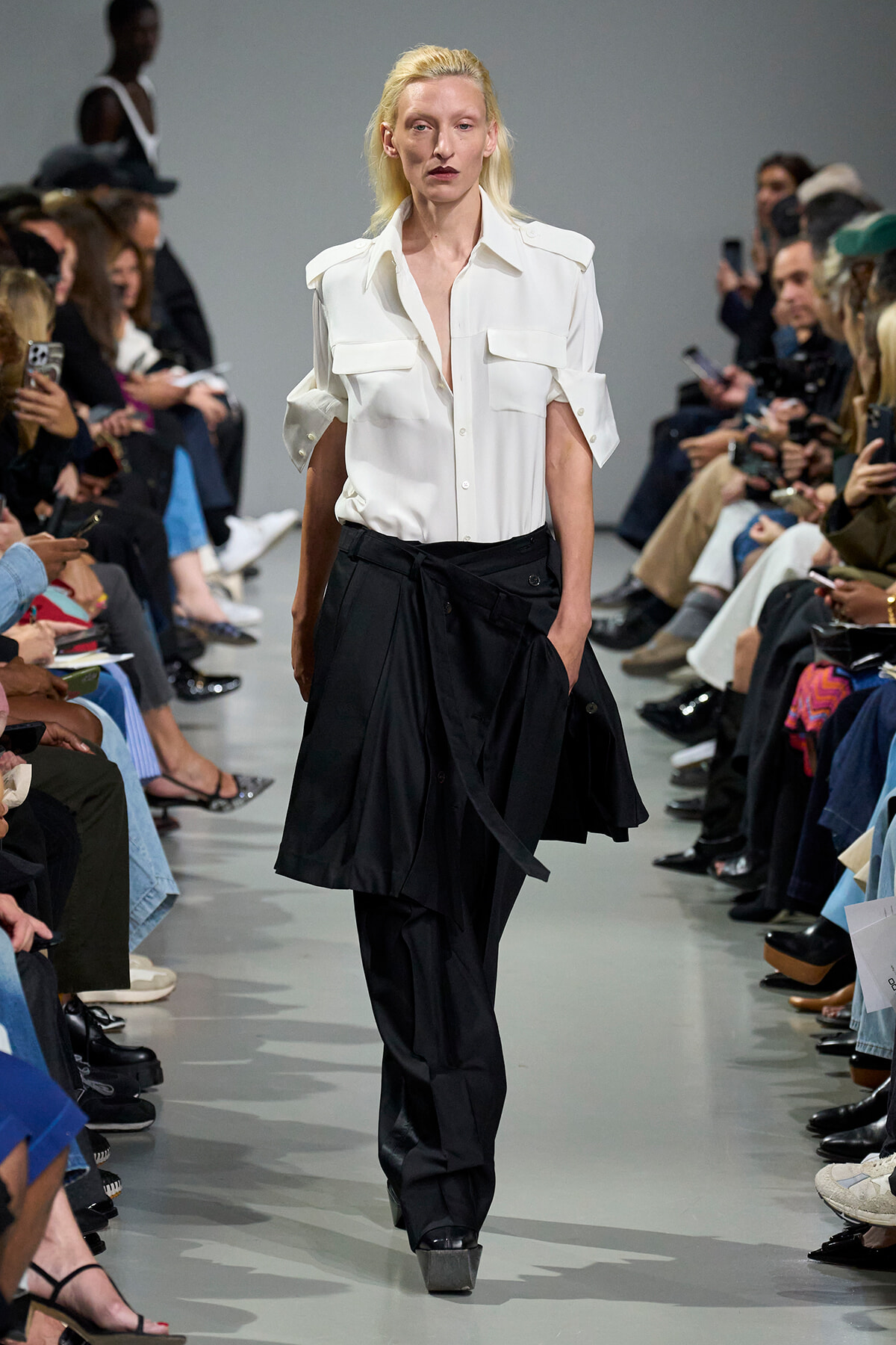 Fashion model walking a runway in a white button-down blouse and black wide-leg pants, audience watching.