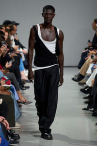 Male model on a runway wearing a black and white layered tank top and loose black pants, audience on both sides.