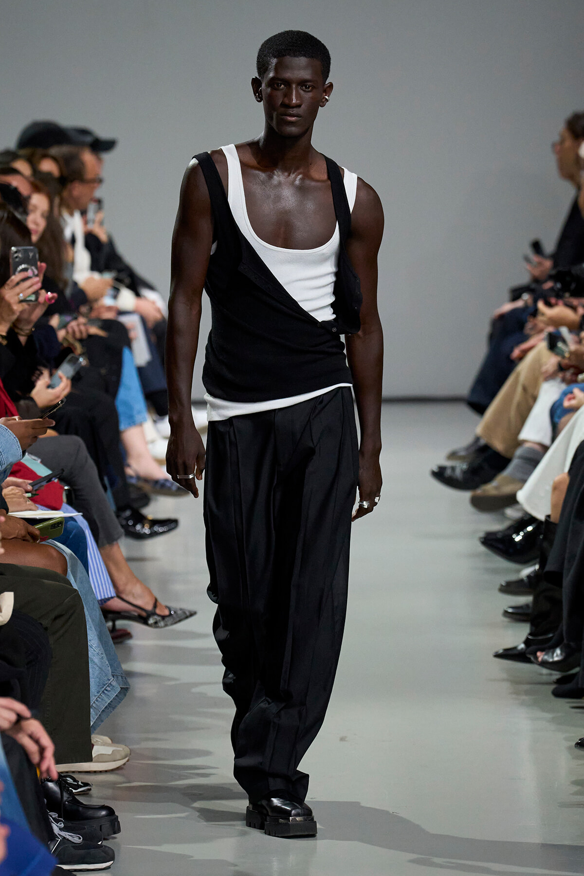 Male model on a runway wearing a black and white layered tank top and loose black pants, audience on both sides.