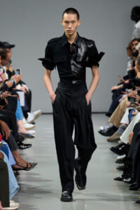 Male model walks a fashion runway in a black leather short-sleeve shirt and matching high-waisted pants, audience on both sides.