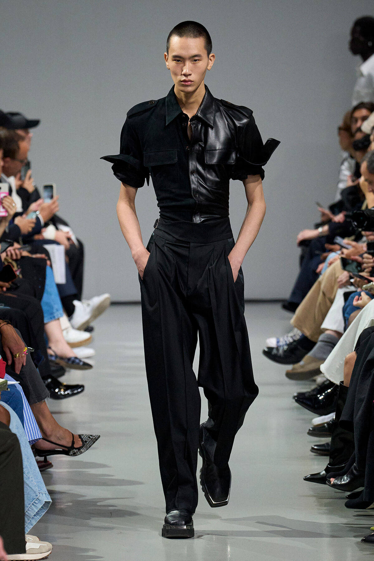 Male model walks a fashion runway in a black leather short-sleeve shirt and matching high-waisted pants, audience on both sides.