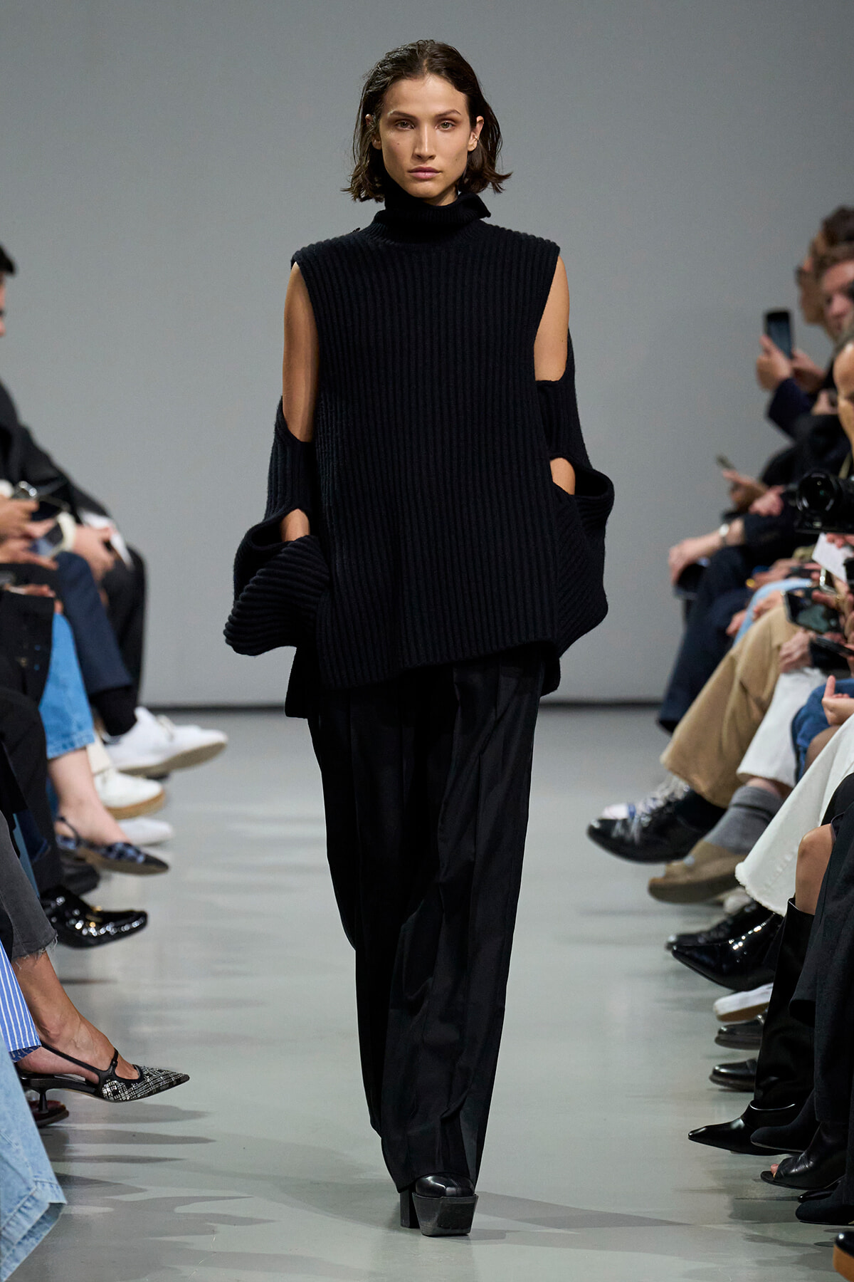Model on a runway wearing an all-black, ribbed sleeveless turtleneck with shoulder cut-outs and wide-leg pants.