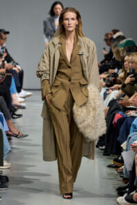 Model walks a fashion runway in a tan coordinated suit with an oversized trench coat; she carries a large cream shaggy bag on her left side while audience watches.
