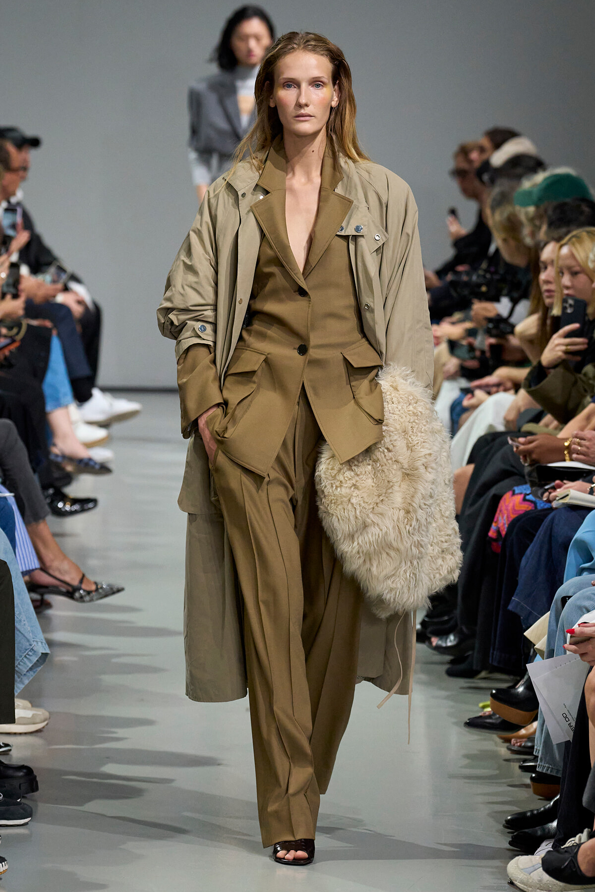 Model walks a fashion runway in a tan coordinated suit with an oversized trench coat; she carries a large cream shaggy bag on her left side while audience watches.