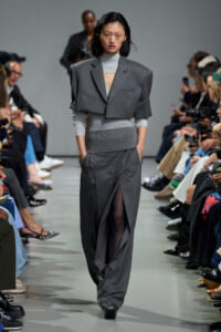 Model walks the runway in a gray tailored suit with a cropped blazer over a light knit top, audience seated on both sides of the catwalk.