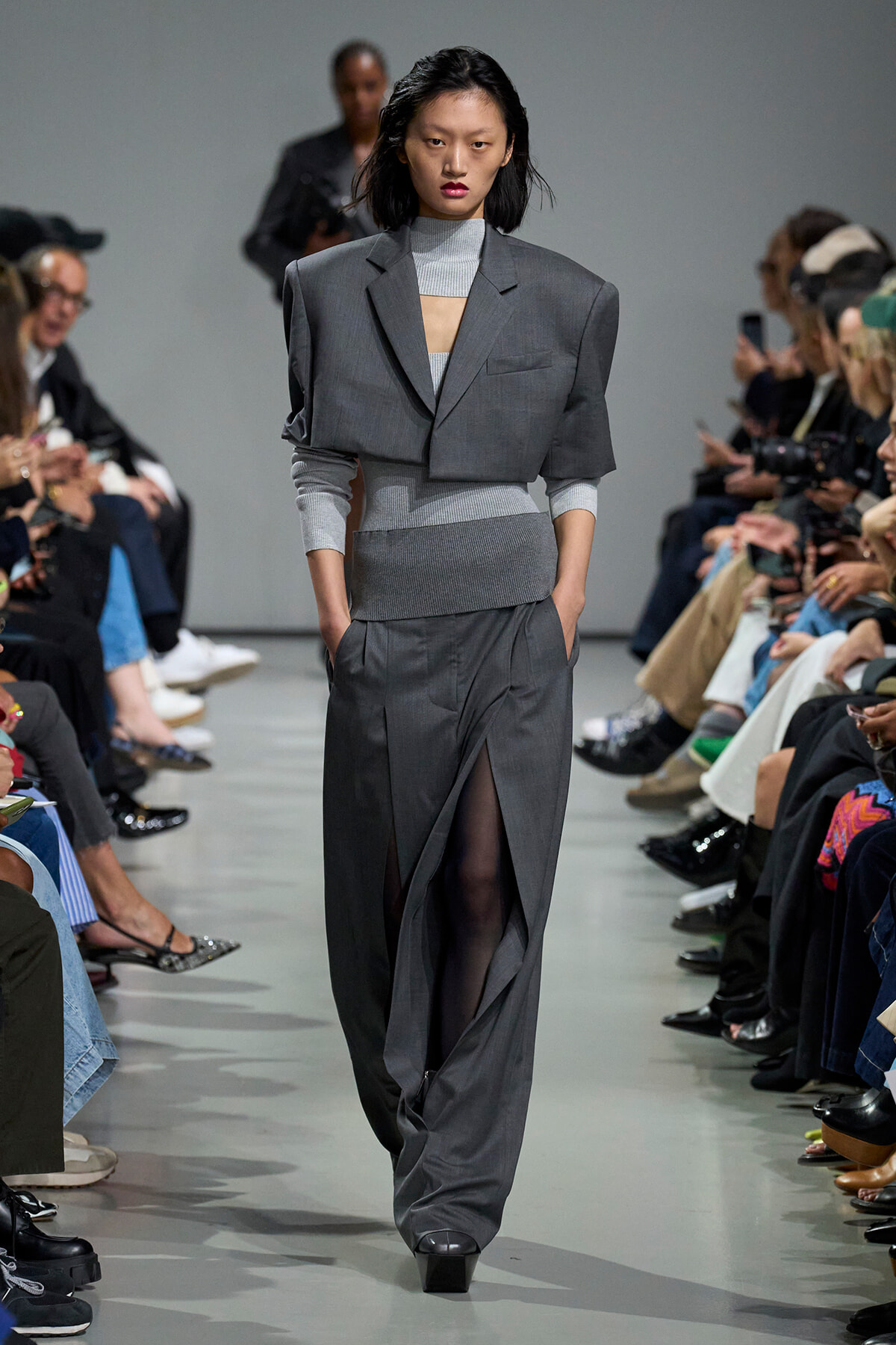 Model walks the runway in a gray tailored suit with a cropped blazer over a light knit top, audience seated on both sides of the catwalk.
