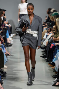 Runway model in a gray suit with blazer and matching shorts, carrying a black handbag as photographers and audience watch.